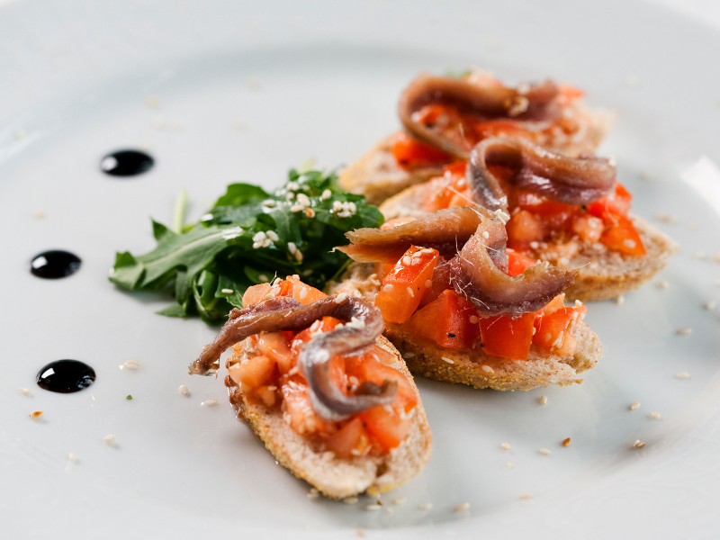 Four Anchovies and diced tomato toast crostini with arugula side on white plate.Deliberico