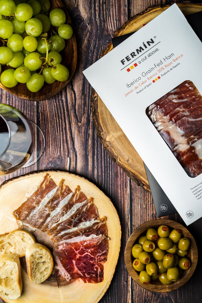 Iberico grain fed ham on wood plates white grapes and olives next to white package by Fermin. Deliberico