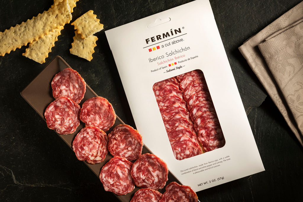 Iberico Salchichon Salami sliced on a dark plate with crackers  and white Packaging by Fermin. Deliberico