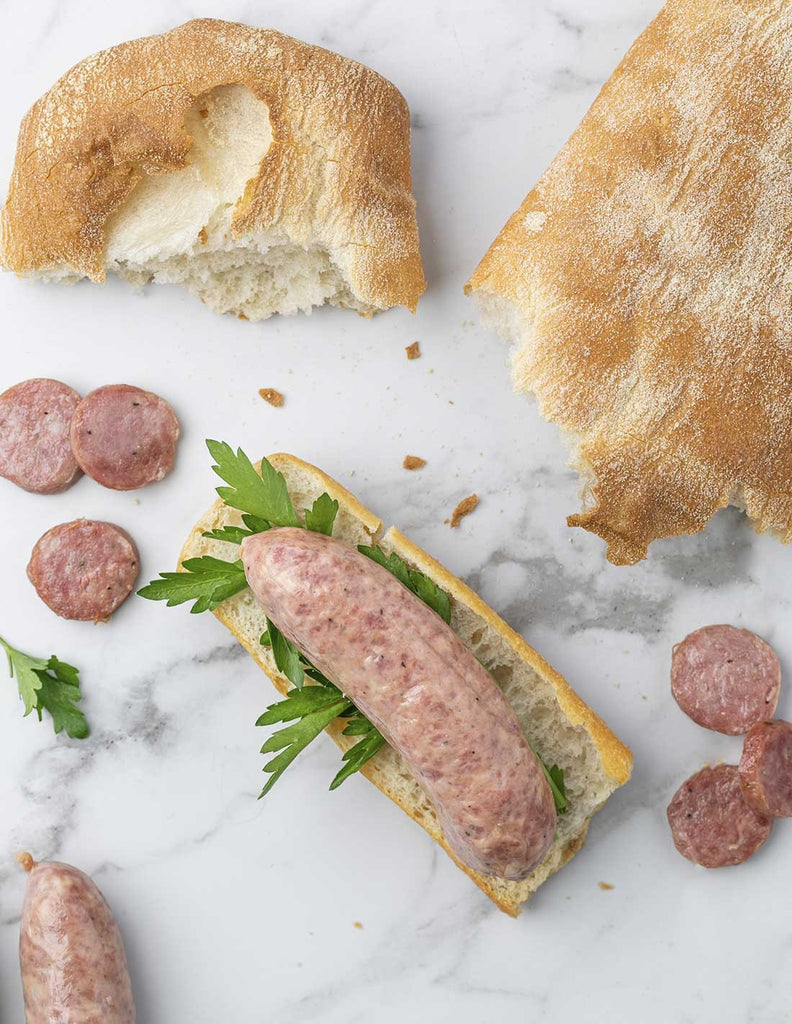 Flat lay of butifarra sausage over a piece of bread and parsey nest to a loaf of bread and slices butifarra on a marble countertop.