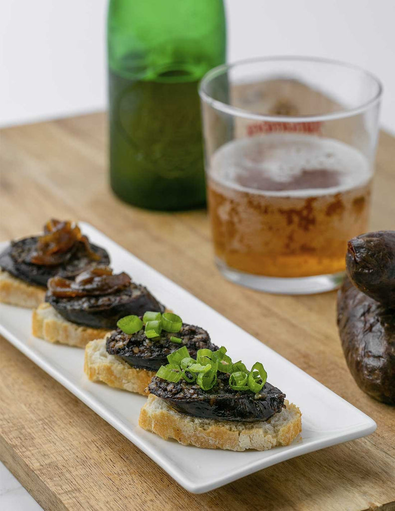 Four Onion Black Sausage slices on bread slices on a long white plate with a green bottle and a glass of beer on a wood board by Dona Juana. Deliberico 
