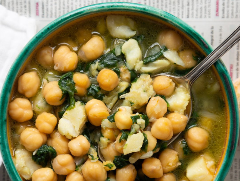 Chickpeas, spinach and cod stew in a green bowl with a spoon over a newspaper background
