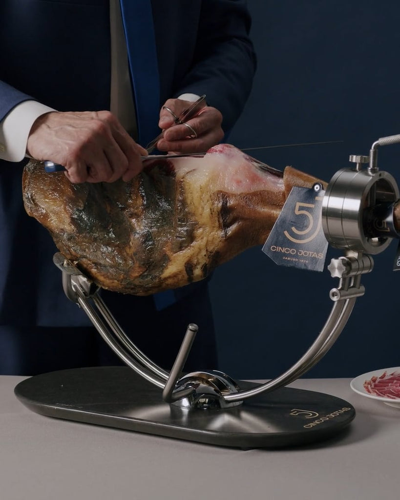 Person cutting into a cured ham on a metal stand with 'Cinco Jotas' branding.