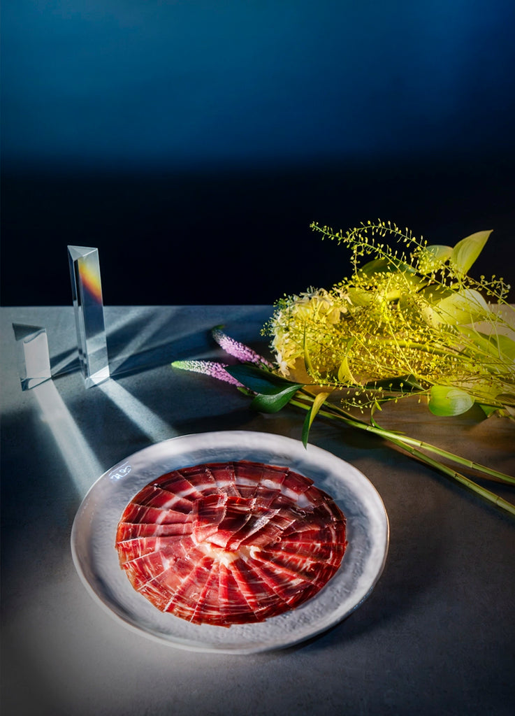 Cinco Jotas Iberico cured ham on a white plate with a dark background and decorative elements.