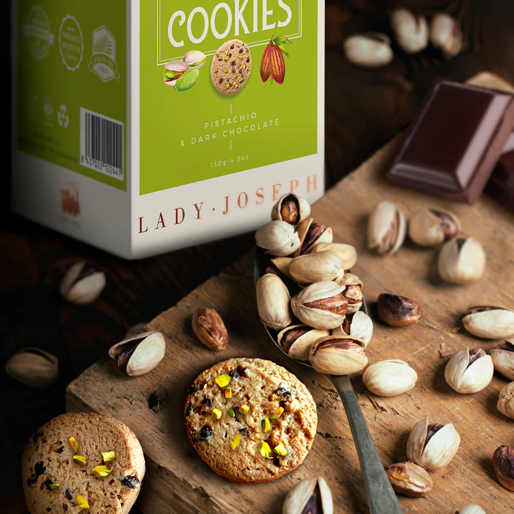 Dark chocolate and pistachio vegan cookies by Lady Joseph in scene with cookies, pistachios and chocolate with a partial view of the box