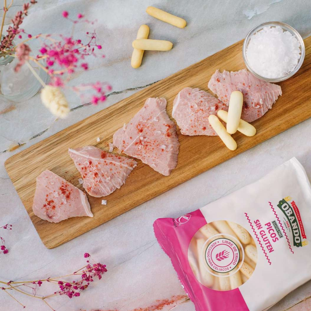 Raw fish fillets on a wooden board with gluten free breadsticks by Obando and a package of breadsticks on a decorative surface.