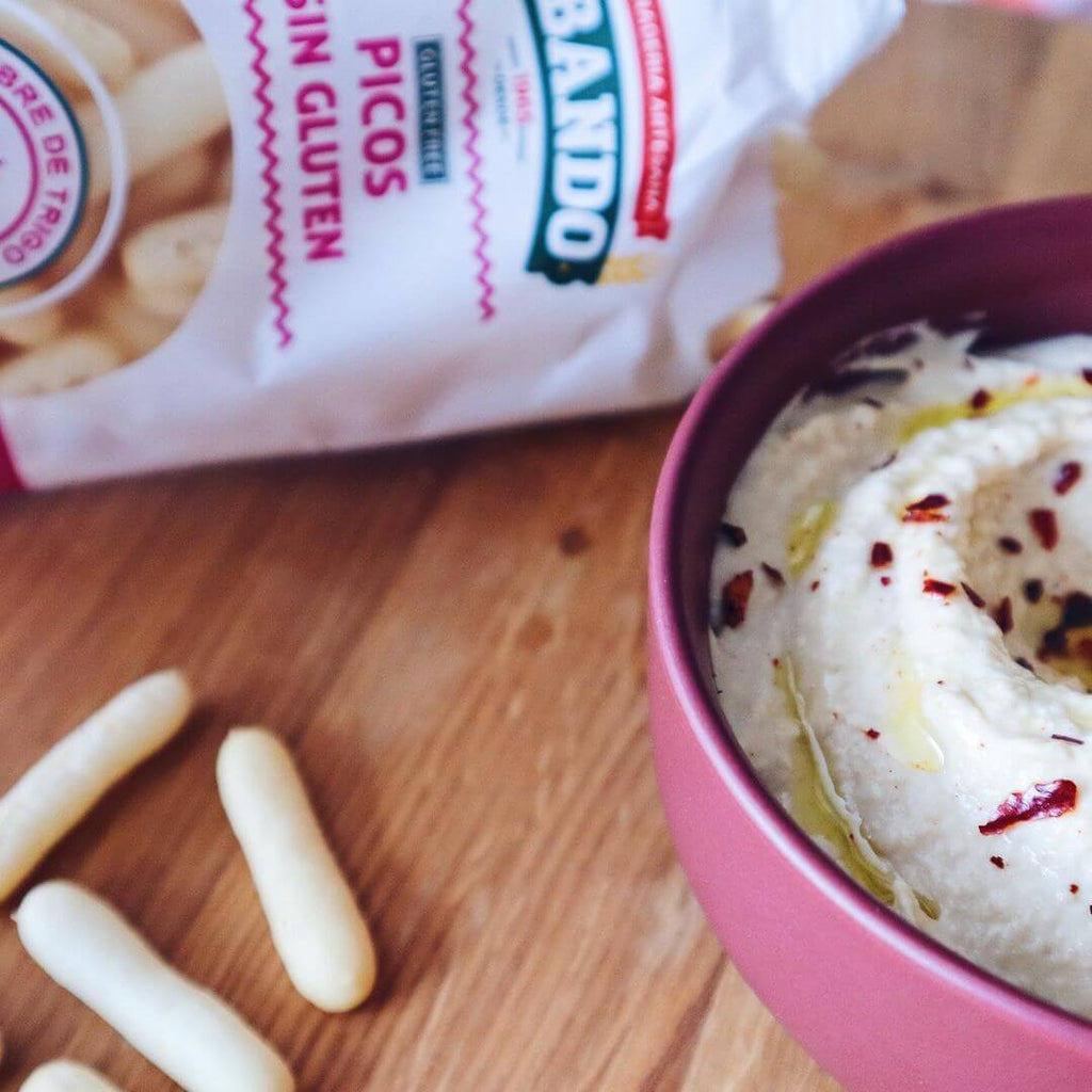 Bowl of hummus with gluten free breadsticks by Obando on a wooden surface, featuring also a Obando Picos gluten-free breadsticks packaging.
