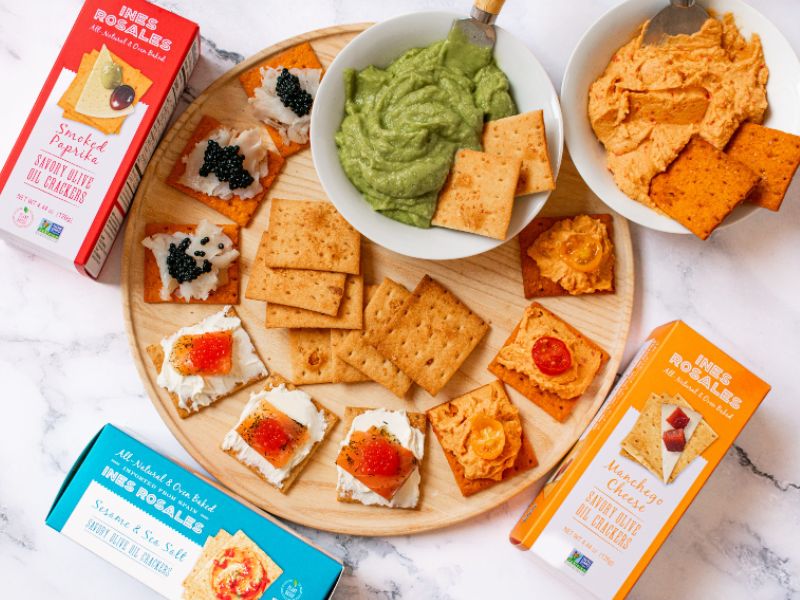 Ines Rosales Crackers Spread with guacamole, hummus, cream cheese over a wood board 