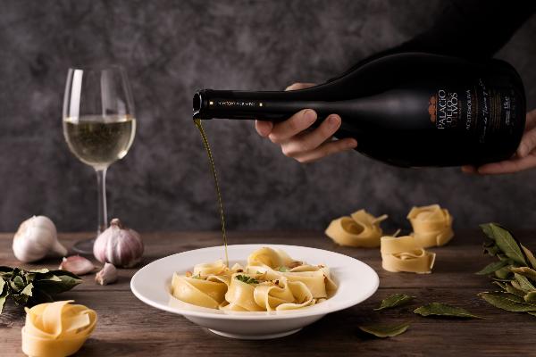 scene of the Palacio de Los Olivos EVOO Magnum bottle pouring olive oil on a pasta dish next to garlic, bay leaves and a glass of white whine on a wood table and a grey background
