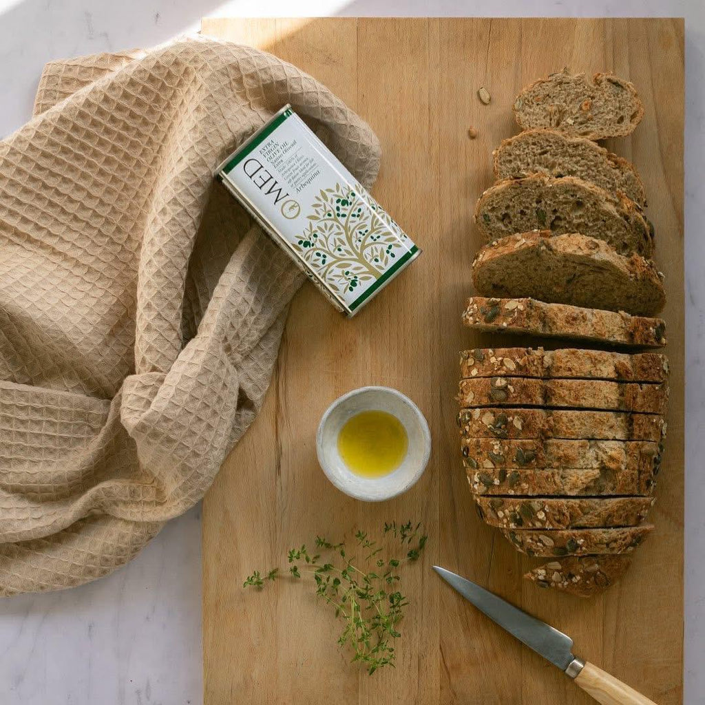 Sliced bread on a wooden board with a towel, O-med Extra Virgin Olive Oil arbequina container, and knife.