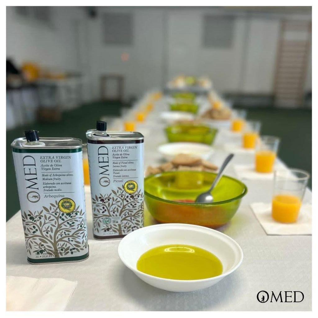 Two bottles of O-med Extra Vigin Olive Oil oon a table with a bowl of oil and snacks in the background as tasting scene