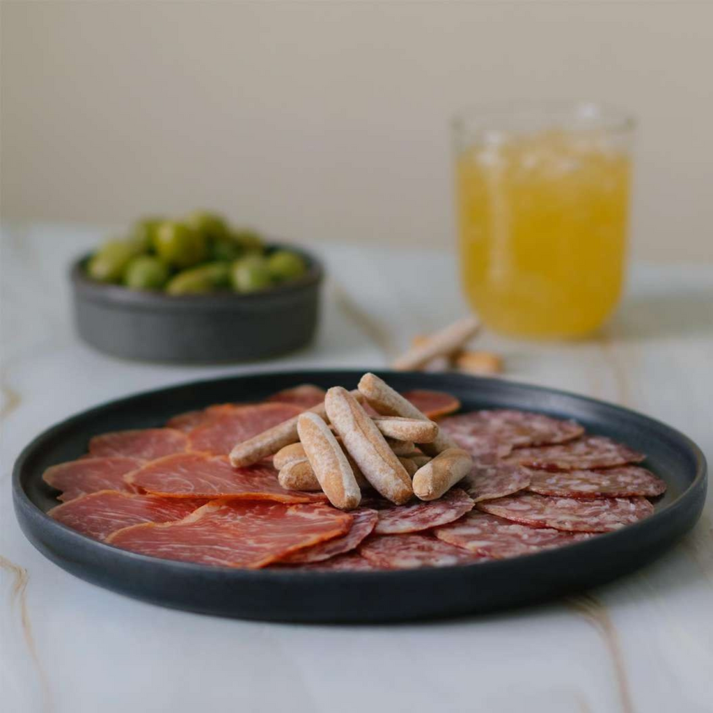 Platter of sliced meats and obando rustic breadsticks with a glass of orange juice on a marble surface.