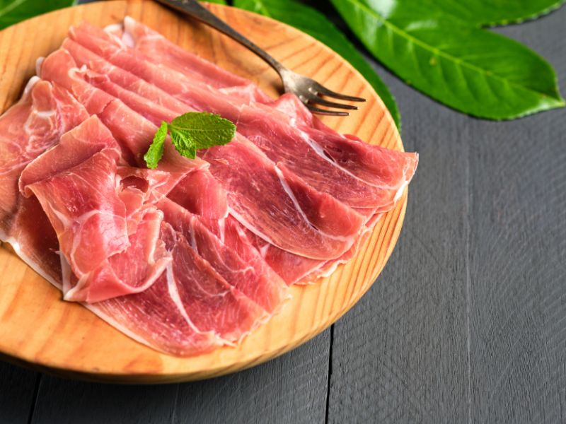 Jamon Serrano sliced on a wooden plate with a fork, on a dark surface with green leaves.