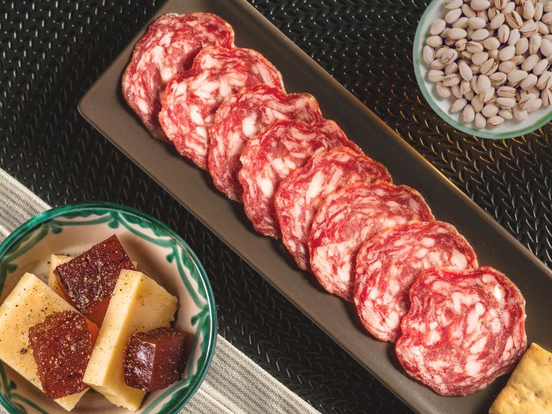 A selection of Spanish tapas including sliced Ibérico Salami, Truffle pistacchio, and Manchego Cheese, presented on a black plate with quince paste and nuts in the background.