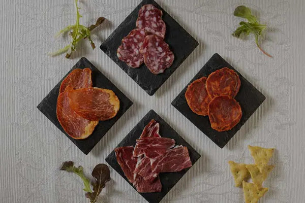 Assorted Fermin iberico cured meats on black slate plates with herbs and crackers on a light background
