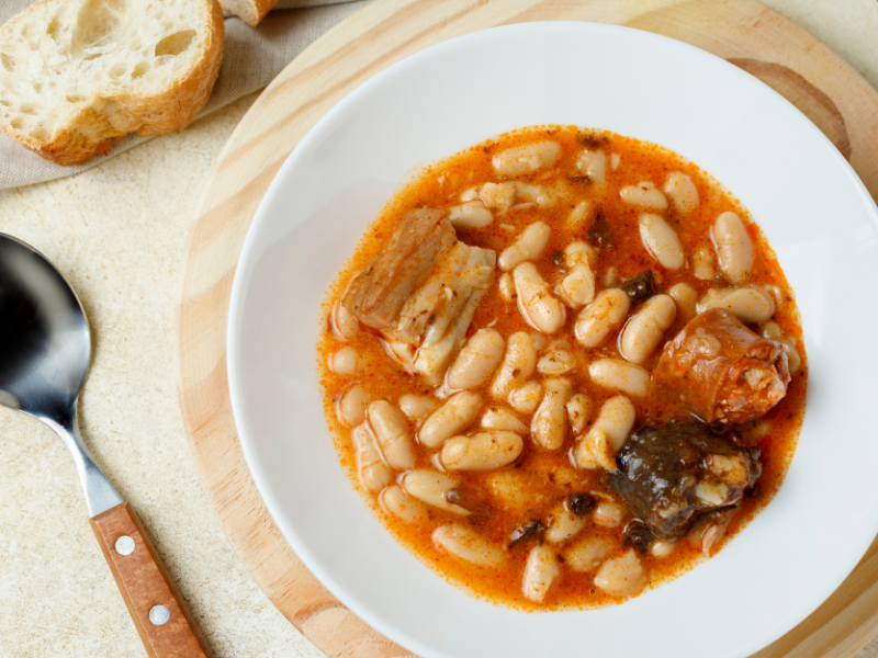 Classic Spanish Fabada white beans dish with chorizo, meat and black sausage in a white plate over a wooden charger and wooden spoon. Deliberico