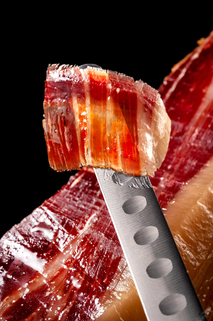 Shiny Iberico ham beautifully cut piece hold with a ham knife on black background by Fermin. Deliberico