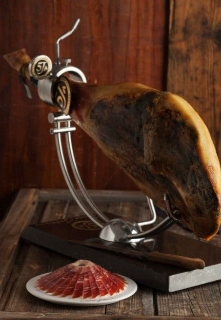 Acorn-fed 100% Iberico Ham on ham holder next to a plate of sliced ham with a wood background by Cinco Jotas. Deliberico