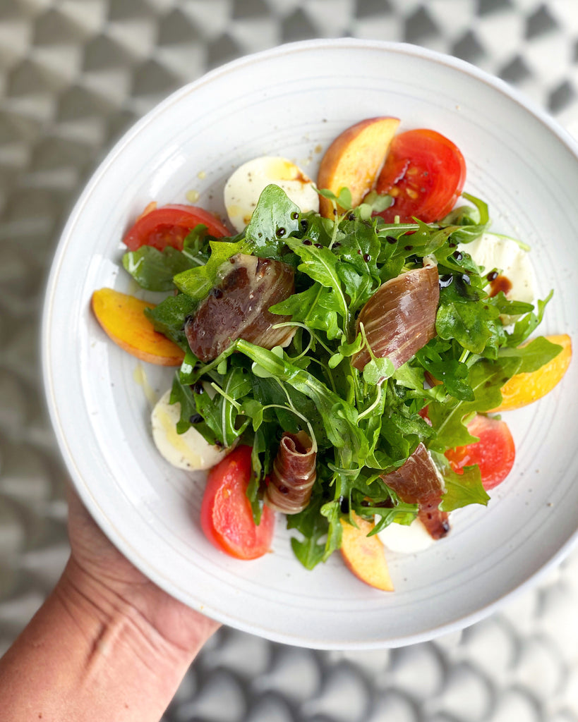 Arugula, tomatoes and iberico ham salad on a white plate hold by a hand by Fermin. Deliberico
