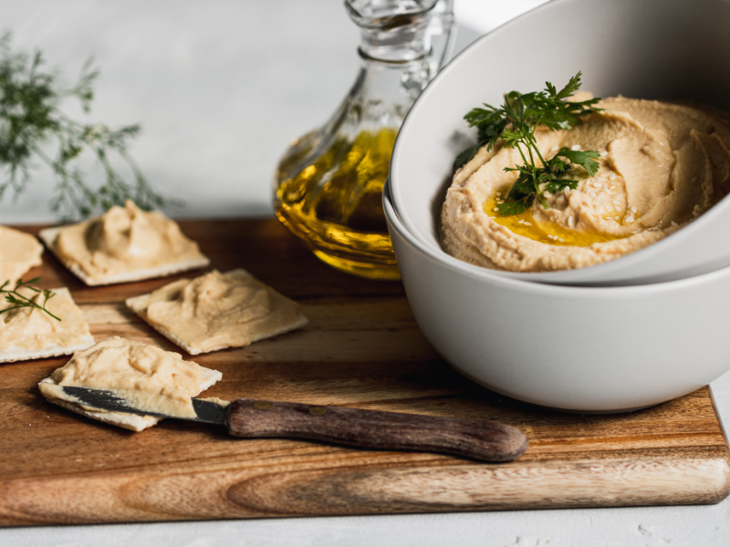 Crackers with cream like hummus on a bowl over a wood board with a knife and olive oil. Deliberico