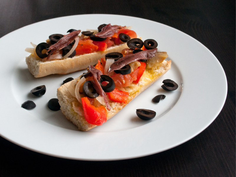 Tapa of bread sliced with pepper strips, sliced black olives and anchovies on a round white plate and a black background. Deliberico