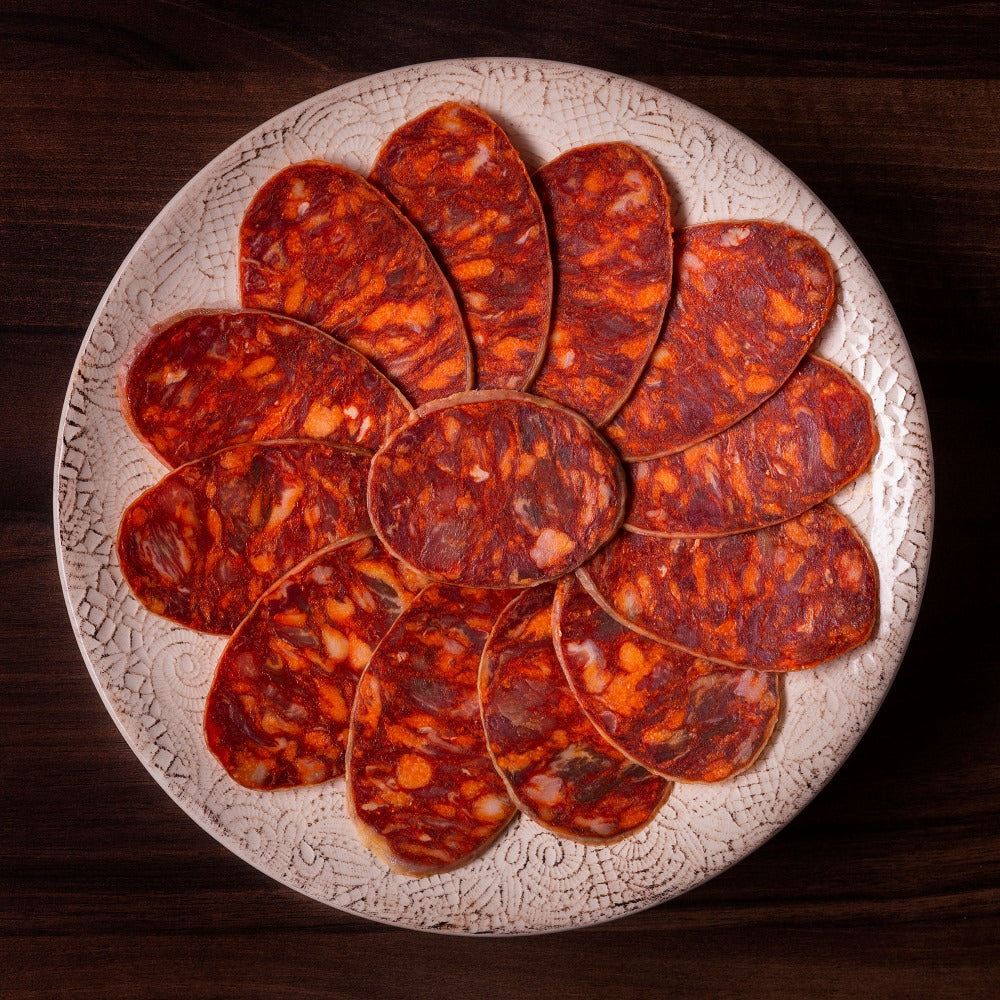Iberico 100 Acorn-Fed Chorizo sliced on white plate with wood background by Fermin. Deliberico