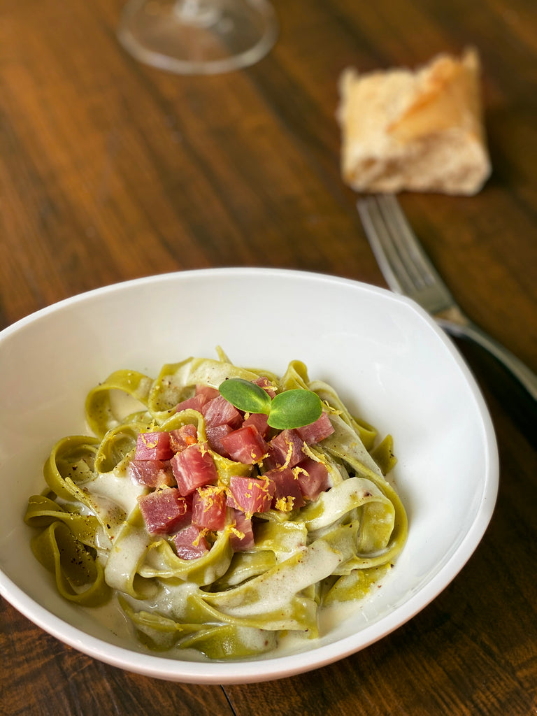 Green pasta with serrano loin topping in a white bowl on wood table by Fermin. Deliberico
