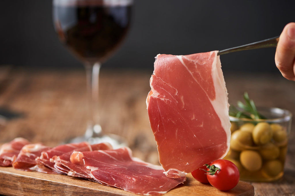 Serrano ham slices on a wood board with olives, tomatoes and red whine glass by Fermin. Deliberico