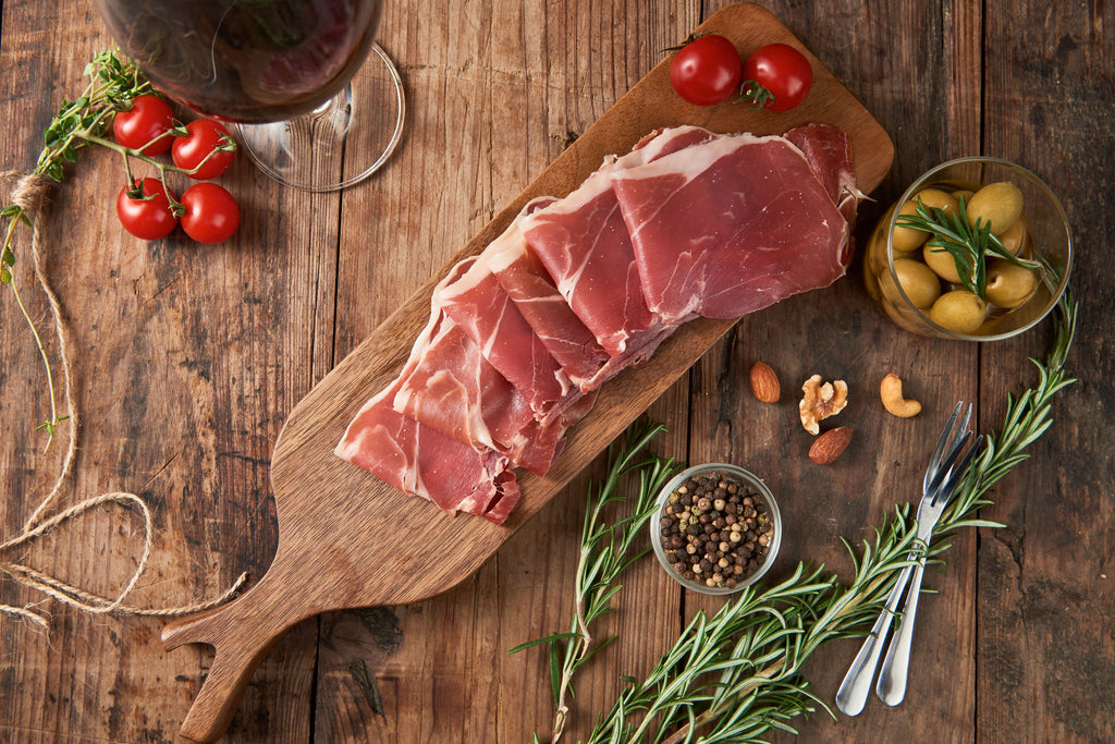 Serrano ham slices on a wood board with olives, tomatoes, nuts and rosemary by Fermin. Deliberico