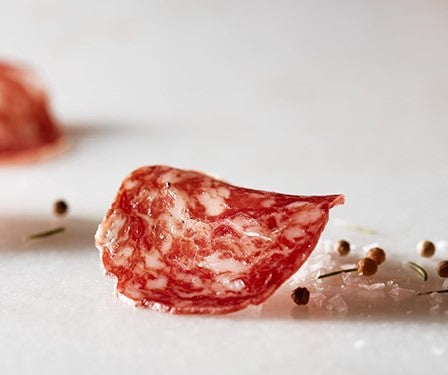 Thin Salchichon salami with pepper and rosemary leaves on a marble surface by Marcos Salamanca. Deliberico
