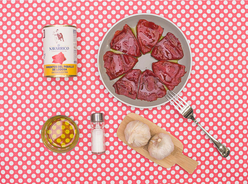 Flat lay of whole piquillo peppers on white plate, olive oil, salt, garlic on wood tray on polka dot white and red background by El Navarrico. Deliberico