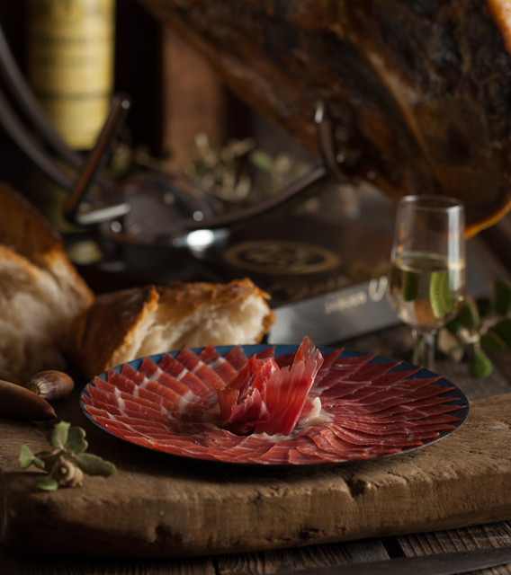 Acorn-fed 100% Iberico Ham sliced on a blue plate with bread and a white wine glass with a ham in the background by Cinco Jotas. Deliberico