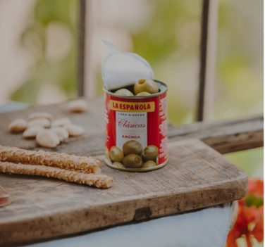 La ESpanola Anchovies Stuffed Olives mini can on wood board with breadsticks and almonds. Deliberico