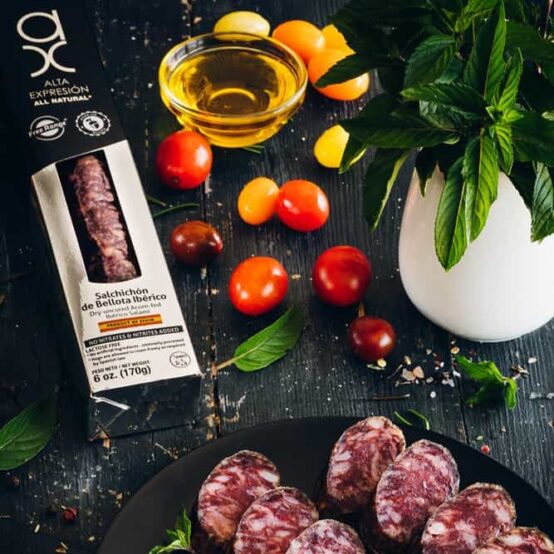 All Natural 100% Iberico Salami Iberico Acorn-Fed in black plate with tomatoes, olive oil and parsley and black and white box by COVAP. Deliberico